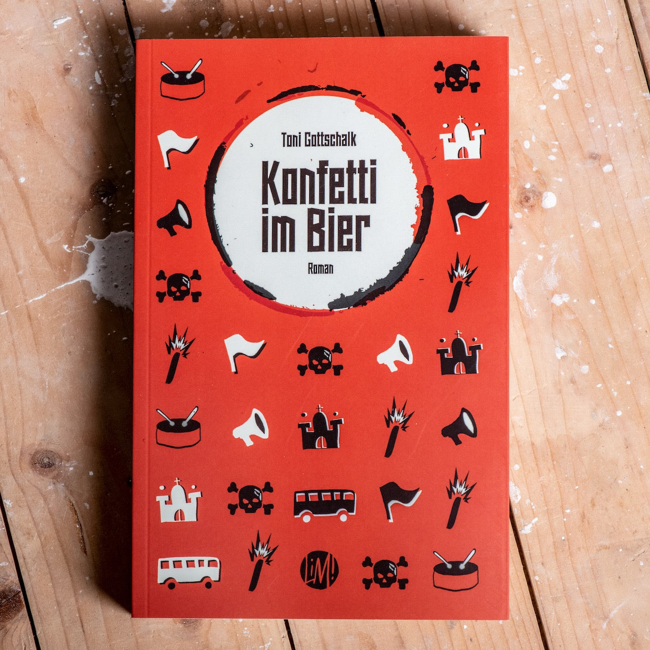 Auf dem Bild sieht man einen Holzfußboden. Auf diesem liegt ein rotes Buch auf dem steht Toni Gottschalk Konfetti im Bier Roman. Es befinden sich außerdem viele, kleine, verschiedene Grafiken auf dem Cover. Diese sind schwarz und weiß. Zum Beispiel Totenköpfe, Busse, Trommeln, Fahnen und mehr.